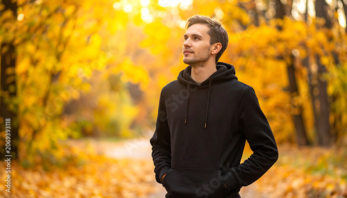 Young man in a black hoodie standing on a path in an autumn forest with golden leaves, looking up thoughtfully, evoking a sense of peace and seasonal change