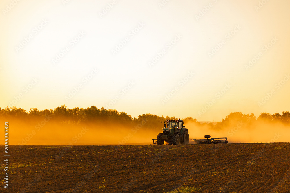 Fototapeta premium Tractor Working in Field with Dust Cloud at Sunset Golden Hour