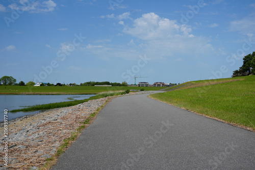 A wide empty asphalted bicycle and walking path running along the banks of the Elbe river near Brunsbüttel, Germany, bordered by a green grassy field on the left and the blue water on the right