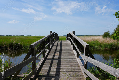 A scenic full shot of a rustic wooden footbridge crossing over a small drainage canal in the rural countryside near Kudensee, Dithmarschen, Germany, on a summer day