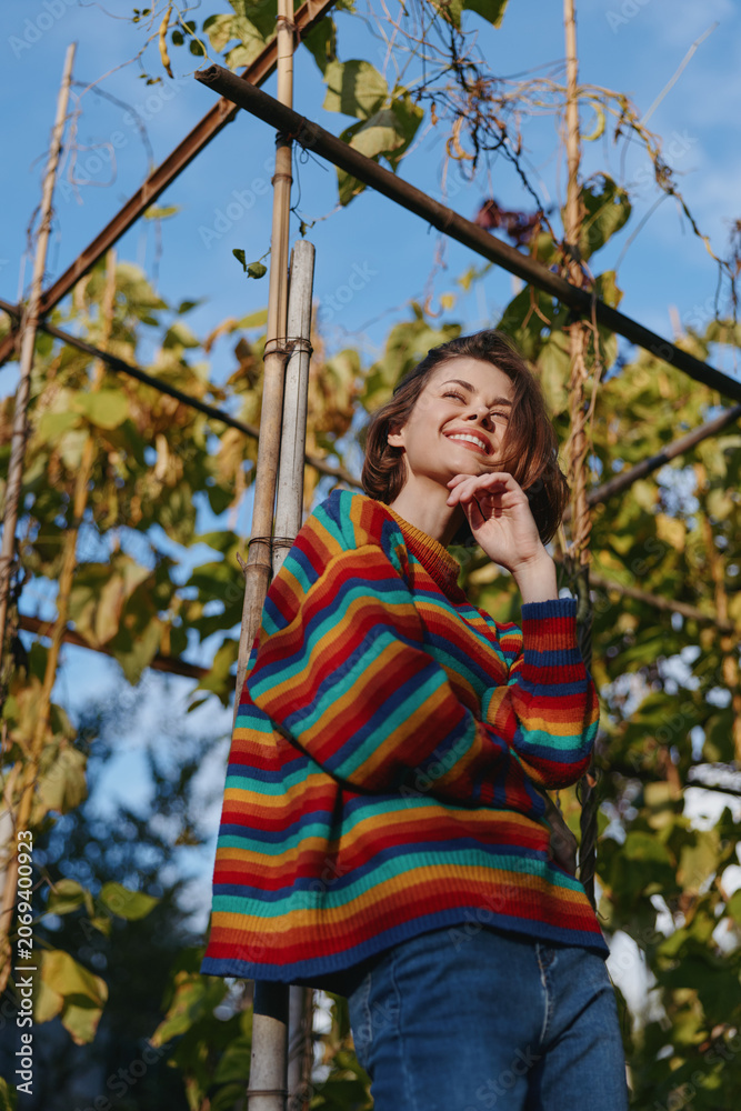 Fototapeta premium Woman in a colorful striped sweater leaning on a bamboo trellis outdoors, smiling and thoughtful in a casual outfit with jeans in an autumn garden lifestyle and fashion portrait.