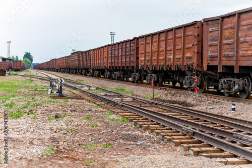 A long industrial freight train consisting of rusty brown open high-sided wagons stands on the tracks of a sorting yard under a cloudy sky. 