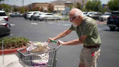 An elderly man pushes a shopping cart filled with groceries through a parking lot after shopping at a grocery store