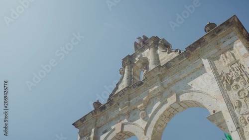 Aerial drone shot looking up at the Zapopan Arch, capturing its decorative columns and eagle sculpture at the top against a vivid blue sky in Guadalajara, Jalisco