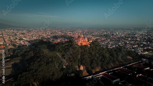 Aerial drone orbits the hilltop church atop the Cholula pyramid at dawn, with the Puebla skyline spreading below and the Popocatepetl and Iztaccihuatl volcanoes rising on the horizon