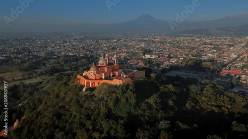 Aerial drone orbits the hilltop church atop the Cholula pyramid at dawn, with the Puebla skyline spreading below and the Popocatepetl and Iztaccihuatl volcanoes rising on the horizon