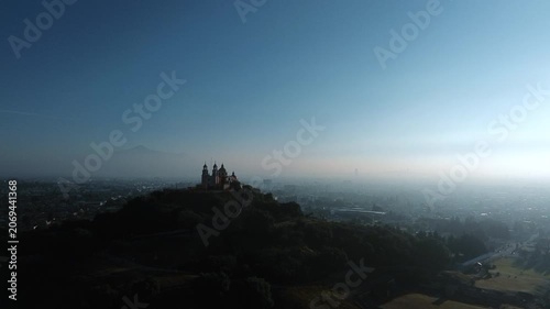 Aerial drone orbits the hilltop church atop the Cholula pyramid at dawn, with the Puebla skyline spreading below and the Popocatepetl and Iztaccihuatl volcanoes rising on the horizon