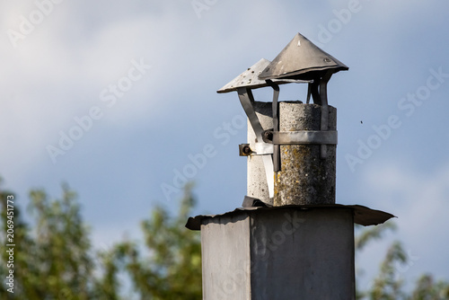 A weathered metal chimney cap with bolts and seams catches warm light against a soft sky showing practical design texture and rural utility atop a simple rooftop vent and flue