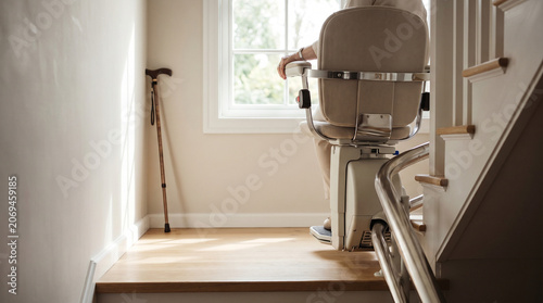 Elderly Mobility Aid: A person is using a stairlift to ascend a set of indoor stairs, with a walking cane leaning against the wall nearby, symbolizing assistance and independence for seniors.
