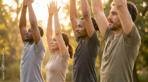A diverse group of adults practicing yoga outdoors in a park during a sunny day, with their arms raised in a meditative pose.