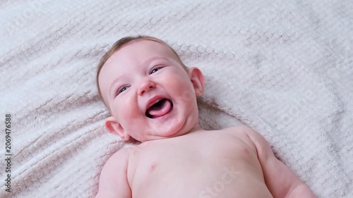 Laughing baby lying on soft white blanket. Joyful infant child showing toothless smile and playing. Excited toddler discovery and development concept. Innocent childhood emotion.