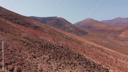 Drone footage flying above a rocky volcanic mountain slope in Fuerteventura, Canary Islands, Spain. Aerial view of arid terrain, red volcanic soil and rugged hills in the island interior.