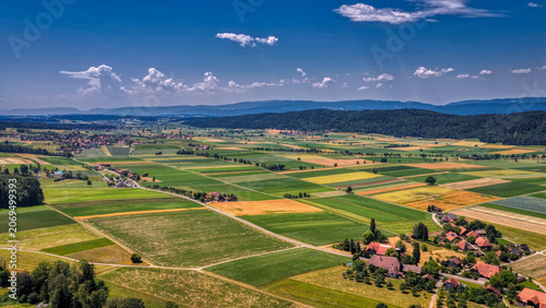 Panoramische Luftaufnahme einer ländlichen Agrarlandschaft mit bunten Feldern, Bauernhöfen, Dörfern und Waldgebieten unter blauem Himmel mit Sonnenstrahlen