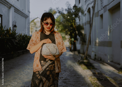 Fashionable adult Asian woman in sunglasses and layered outfit with batik bottom opening a small her bag standing on alley city street during sunsets.