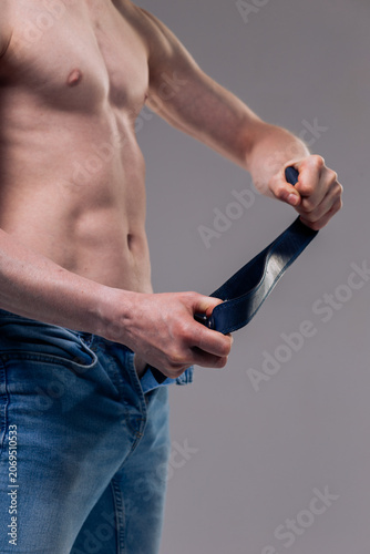 Bild auf Leinwand A muscular man in blue classic jeans with a bare torso is holding a belt