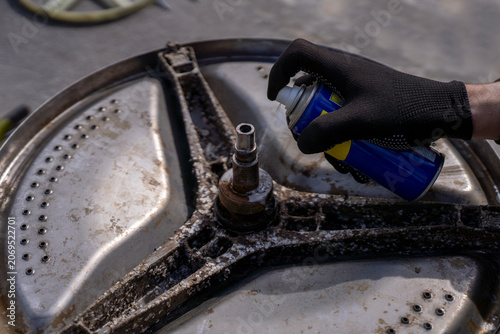 Fotografi Technician hand spraying penetrating lubricant lubricant onto a rusty spider arm bracket shaft of a disassembled stainless steel washing machine drum during appliance repair service