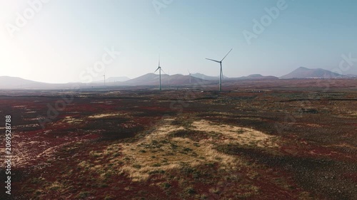 Drone flight over a wind farm in Fuerteventura, Canary Islands, Spain. Multiple wind turbines stand across volcanic terrain with mountains in the background and clear sky.