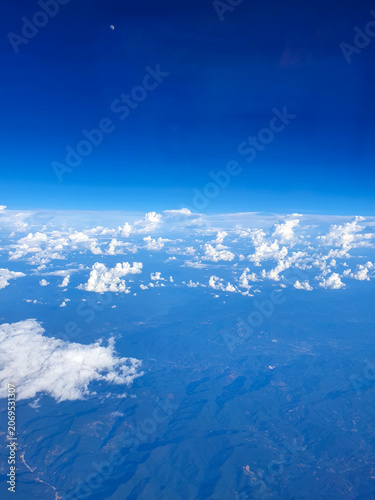 Panoramic aerial view from an airplane window showing towering white cumulus clouds above a mountainous landscape. The deep blue sky fades toward the horizon, creating a dramatic high-altitude scene 