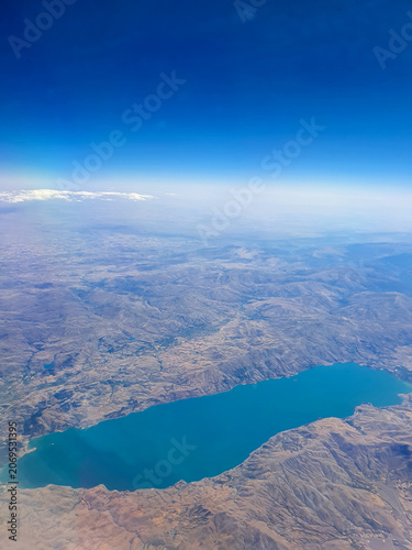 High-altitude aerial view of a blue lake surrounded by rugged arid mountains and dry terrain under a clear blue sky. The landscape is photographed from an aircraft, showing natural landforms and the s