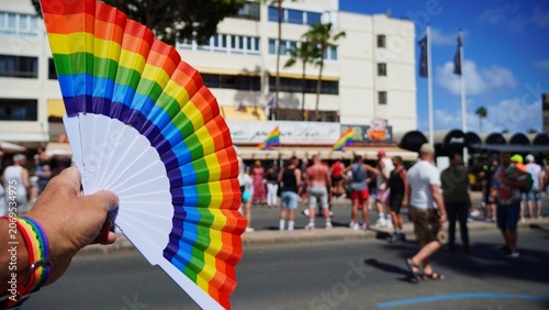 Person's hand holding a vibrant rainbow fan and wearing a matching bracelet, symbolizing diversity, equality, and support during an outdoor LGBTQ+ Pride parade celebration