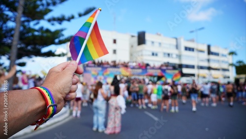 Hand holding a vibrant rainbow flag and wearing a matching bracelet, symbolizing LGBTQ+ pride, diversity, and inclusion amidst a celebrating crowd at a street parade