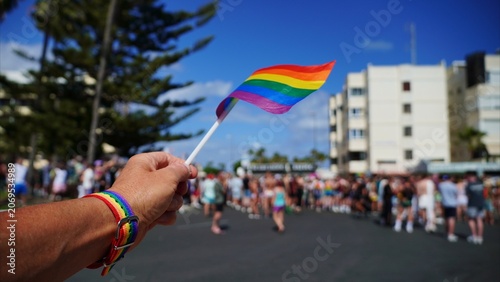 Person's arm with a rainbow wristband holding a small vibrant rainbow flag, symbolizing LGBTQ+ pride, diversity, and support at a large outdoor parade with a celebratory crowd