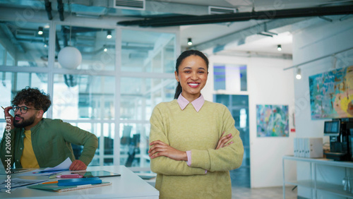 Smiling businesswoman standing with crossed arms. Joyful female looking at camera. Curly Caucasian male coworker discussing project. Creative team working inside contemporary office space.