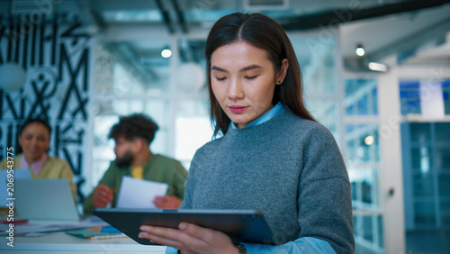 Cheerful Asian businesswoman holding tablet device. Smiling female looking at camera. Curly Caucasian male colleague reviewing papers. Team collaborating inside contemporary startup office.
