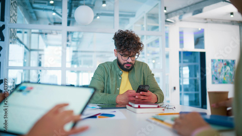 Focused male employee sitting at desk with notebook and tablet. Young man browsing content on smartphone. Worker holding mobile device and reading messages. Business professional working alone.
