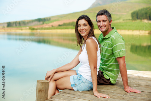 Lake, portrait and relax with happy couple in nature for holiday getaway or natural scenery. Man, woman or travel with smile on lakeside bridge for outdoor weekend, summer vacation or adventure