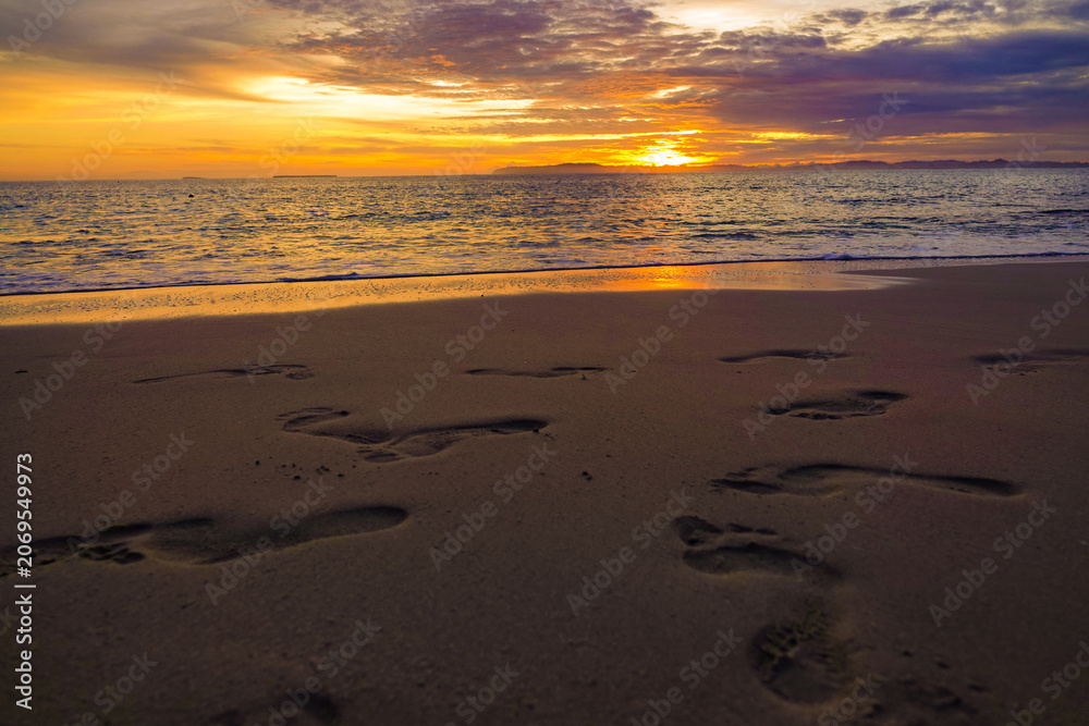 Obraz premium LENS FLARE: Numerous footprints trace paths along wide sandy shoreline under colorful sunset. Vibrant setting sun breaks through clouds and spreads a warm light across ocean tides on an island beach.
