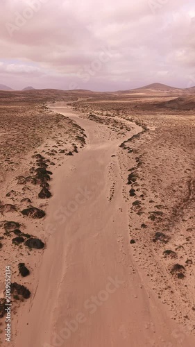 Vertical aerial view of a sandy desert track winding through the barren volcanic landscape of Fuerteventura, Canary Islands, Spain under dramatic cloudy skies.