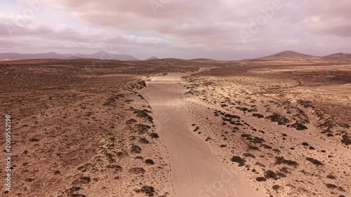 Drone flight above a sandy road crossing the arid volcanic landscape of Fuerteventura, Canary Islands, Spain. Remote desert terrain with mountains and cloudy sky.