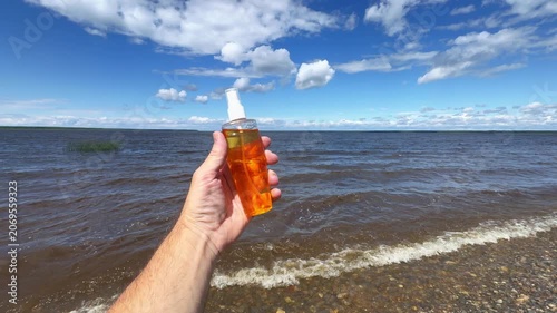 Hand presents tanning lotion spray bottle against lake waves and blue sky. Concept: summer skincare, sun care, travel product and outdoor relaxation.