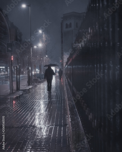 A moody and atmospheric cinematic shot of a wet city street at night during a heavy rainstorm. Blurred streetlights and traffic lights create beautiful neon reflections on the damp, shiny pavement. 
