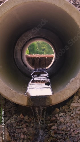 Water flows through a pipe and out into an open area surrounded by trees and stones in bright daylight
