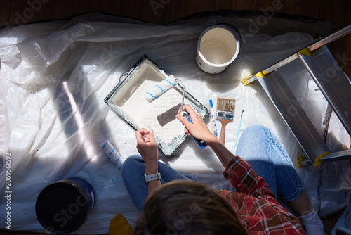 Overhead view of person holding paint roller above tray with bucket, brushes and ladder on protective sheet