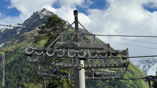 Ski lift system featuring pulleys and cables set against lush green hills and snow-covered mountains under a bright blue sky