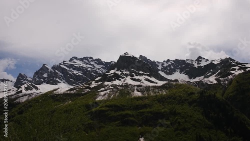Snow-covered peaks rise above vibrant green hills under a cloudy sky, illustrating the stunning contrast of nature's elements in a mountainous region
