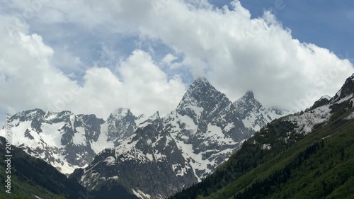 Snow-covered mountain peaks rise dramatically against a cloudy sky, with verdant hills below, capturing the stunning landscape of a mountainous area