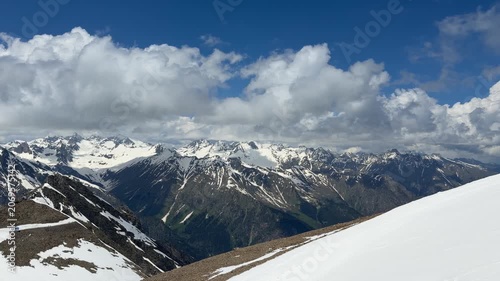 Snow-covered peaks of a mountain range stretch across the horizon under a vibrant blue sky filled with fluffy white clouds