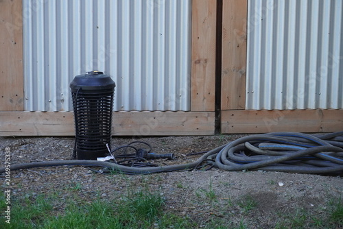 An electronic outdoor bug zapper lantern stands on the ground next to a coiled garden hose against a corrugated metal fence with a wooden frame.