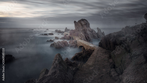 Long exposure of a stone bridge on a rocky coast with misty sea, A breathtaking long exposure landscape photograph of an ancient stone pathway and bridge winding through rugged sea rocks and cliffs.