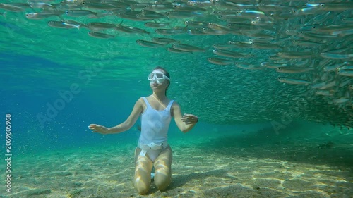 Thousands of Atherina fish create dense, shimmering screen in foreground, girl snorkeler in white freediving gear sits on sandy seafloor and looks at moving silver mass in sun-drenched sandy shallows