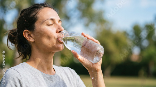 Woman drinking water from glass bottle outdoors. Refreshment during hot summer weather. Female hydrating body to quench thirst. Healthy lifestyle and wellness concept during heat wave.