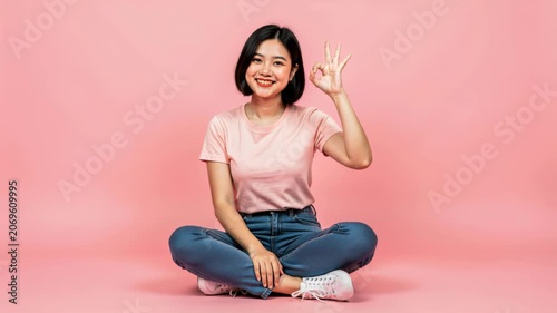 A Woman's Affirmation: A young Asian woman in casual attire sits cross-legged, offering a warm smile and an OK hand gesture, radiating positivity and contentment.