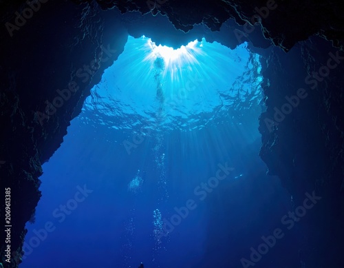 Scuba diver ascending through an underwater sea cave with sunlight streaming through the surface opening, illuminated blue water, ascending air bubbles, dark rocks.