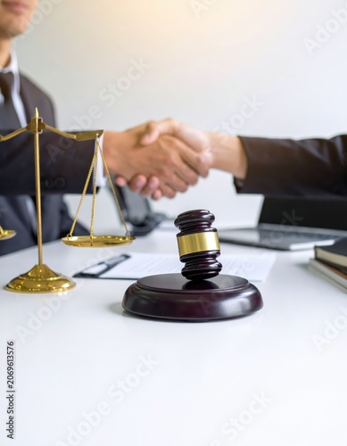 Two business people shaking hands over a legal contract with a wooden gavel and golden scales of justice on a white desk in an office setting.