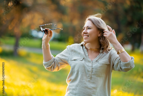 Принти на полотні Smiling woman holding sunglasses and touching hair in summer park
