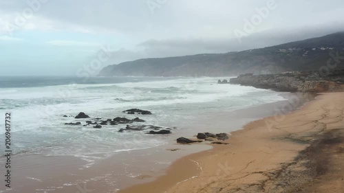 Waves crash against the rocks at Guincho Beach as clouds linger overhead. The golden sand glimmers, revealing nature's power and tranquility after a storm.